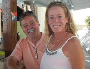 A man and a woman smiling at the camera, standing in a casual outdoor setting. The man has a mustache and wears a peach shirt, while the woman has long hair and wears a white tank top with embellishments.