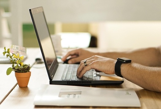 A person wearing a smartwatch types on a laptop at a wooden desk, with a small potted plant and an envelope nearby.