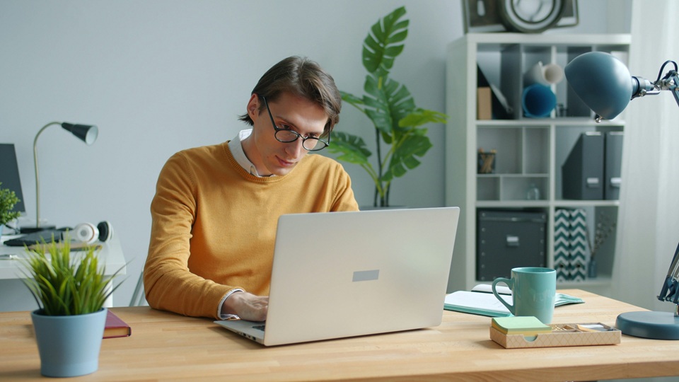 A man wearing glasses and a yellow sweater works on a laptop at a wooden desk in a bright, modern home office with plants, books, a lamp, and a mug.