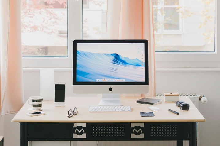 A neat desk with an iMac displaying a wave wallpaper, a wireless keyboard and mouse, a smartphone, a cup of coffee, a notepad, pens, a lamp, and other small items, set in front of a window with peach curtains.