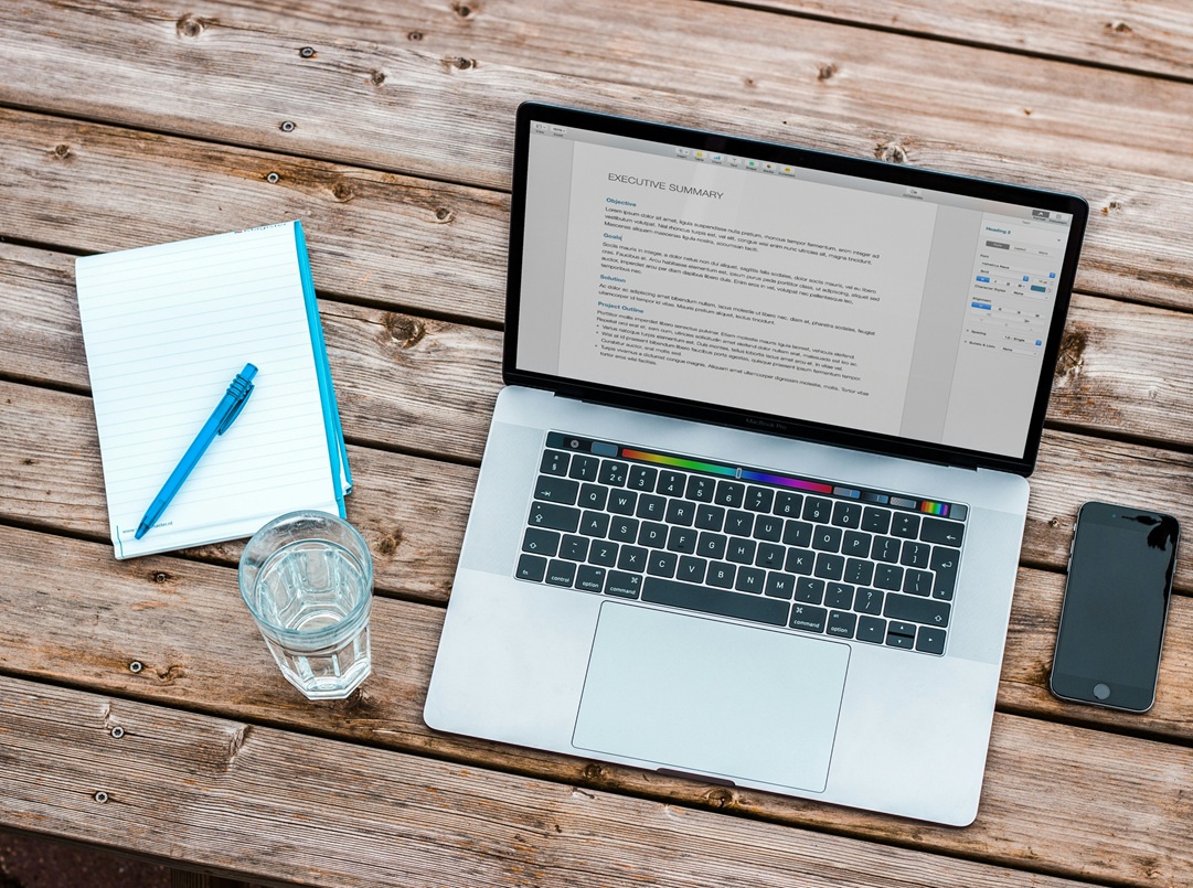 A laptop displaying a document sits on a wooden table next to a notepad with a blue pen, a glass of water, and a smartphone.
