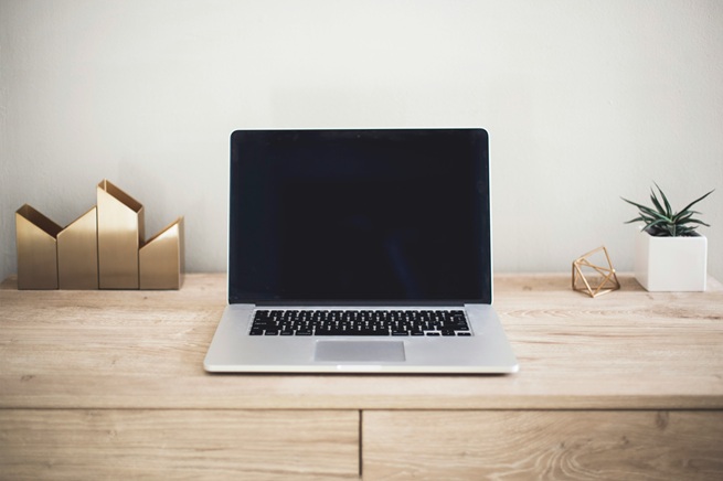 A laptop with a blank screen sits on a wooden desk. To the left are gold geometric bookends, and to the right are a small potted succulent and a gold wire sculpture. The background wall is plain and light-colored.